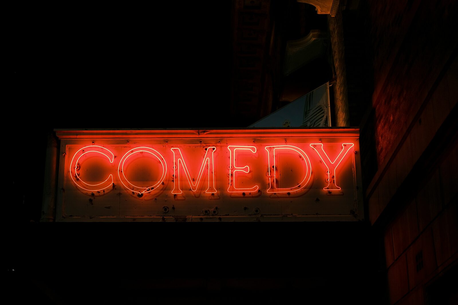 A weathered red neon sign reading COMEDY glowing above a darkened theatre entrance at night.